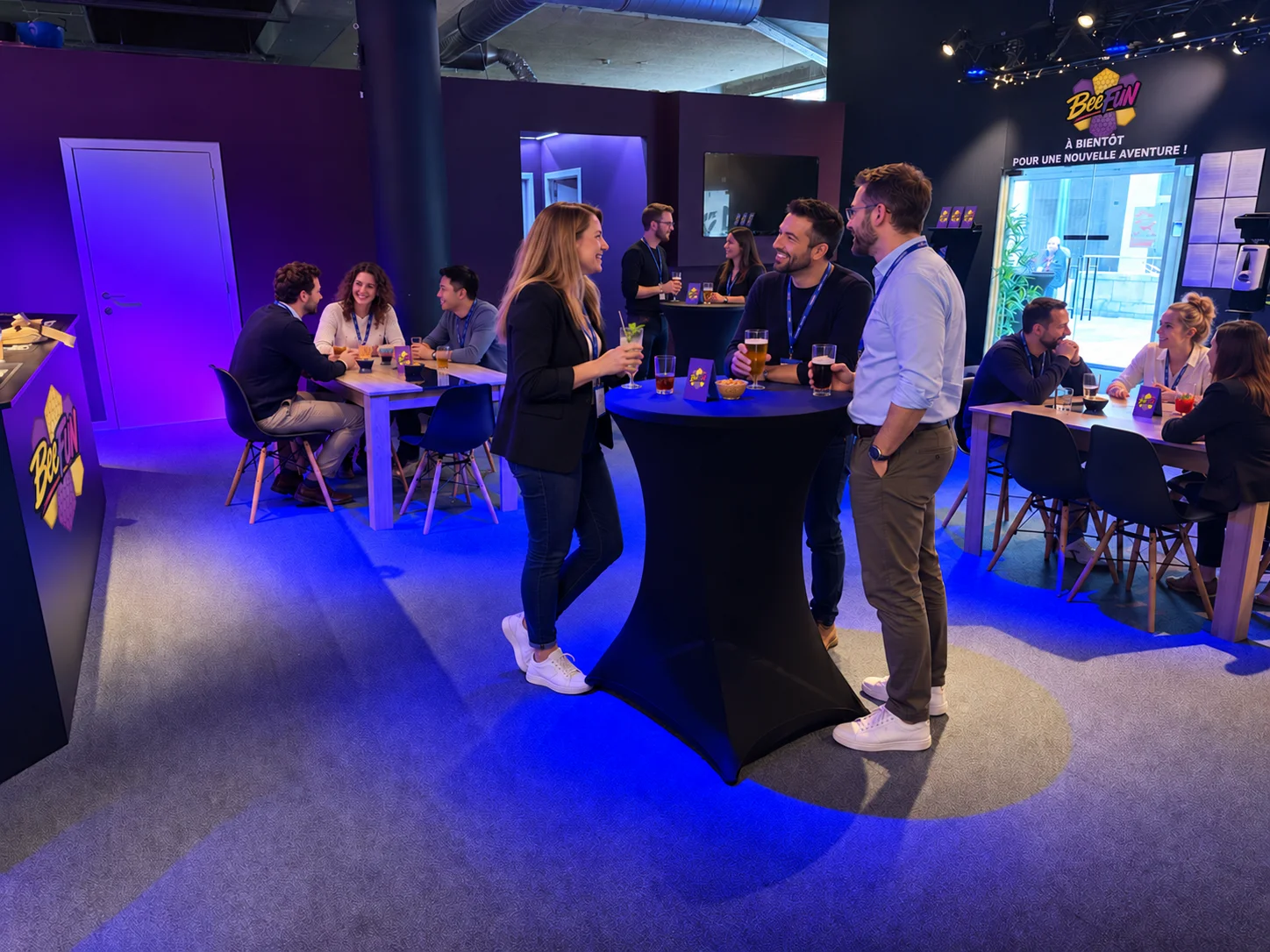 Des personnes échangent autour de tables hautes avec des boissons dans un espace intérieur où d’autres groupes sont assis.